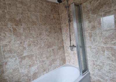a newly tiled beige marbled bathroom wall around a bath with glass screen and overhead shower