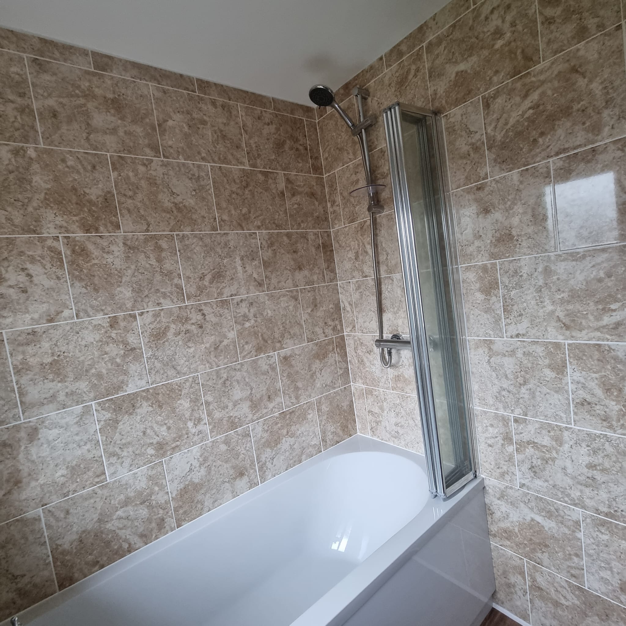 a newly tiled beige marbled bathroom wall around a bath with glass screen and overhead shower