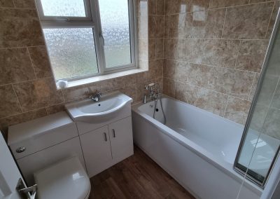 a newly decorated bathroom showing a white toilet, sink and cupboard and bath with a window behind. Beige marble tiled walls and a wooden effect laminate floor