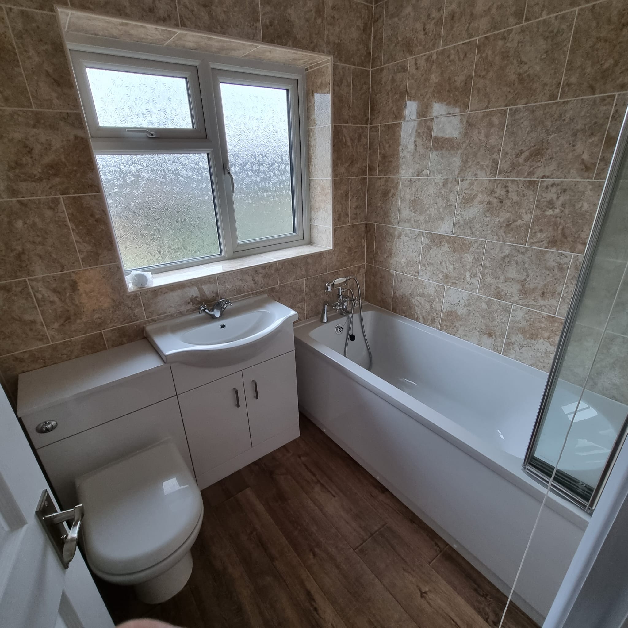 a newly decorated bathroom showing a white toilet, sink and cupboard and bath with a window behind. Beige marble tiled walls and a wooden effect laminate floor
