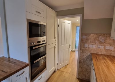 A newly fitted kitchen and hallway with white cupboards, built in oven, wooden worktops and wooden laminate flooring. Olive green paint on the walls.