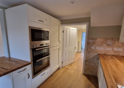 A newly fitted kitchen and hallway with white cupboards, built in oven, wooden worktops and wooden laminate flooring. Olive green paint on the walls.