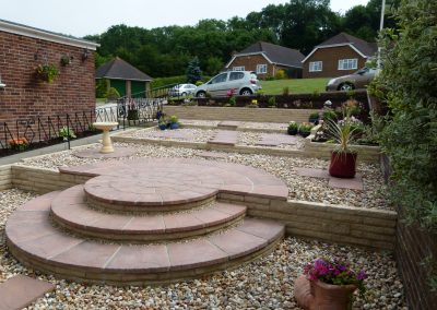 Newly laid front garden area with 3 beautiful circular stepped areas in light paving, gravel tiers and paved areas with containers planted. Light stone walls separate each area