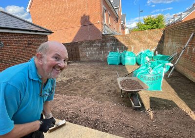 a picture of a smiling man in blue tshirt in excavated garden area with a wheelbarrow and 5 bright green builders bags. Garden surrounded by brick wall