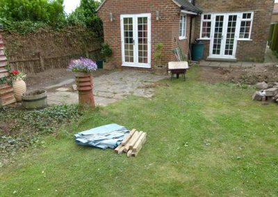 a garden area with old patio and pile of rubble and weeds with builders tarpaulin, wheelbarrow and tools