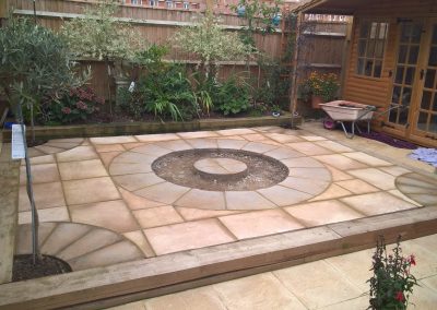 a new patio area with light coloured paving stones, circular designs and a wooden sleeper surround. A new garden border with shrubs in the background and wooden garden building to the side.