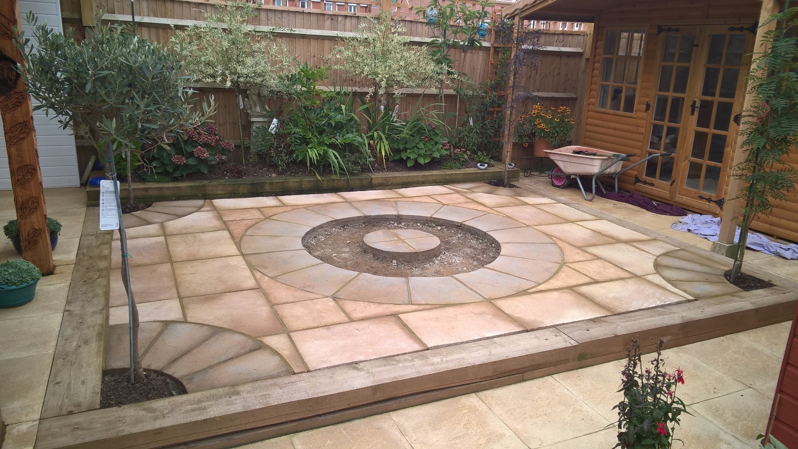 a new patio area with light coloured paving stones, circular designs and a wooden sleeper surround. A new garden border with shrubs in the background and wooden garden building to the side.