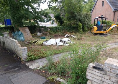 an old garden area full with old path, weeds, builders rubble and a workman on a digger