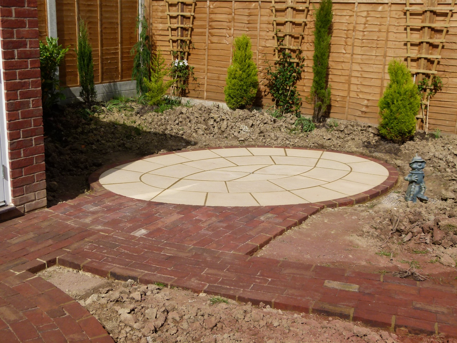 small circular patio with a light stone, surrounded by red brick and newly planted conifers against a fence.