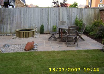 a new patio and small pond area with wooden table and chairs and terracotta garden urn. There is a fence behind and shrubs to the side.