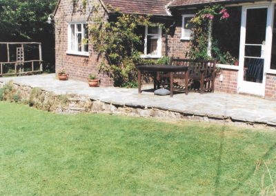 a country style house with climbing roses and a wooden bench and table of a newly laid patio area with brick wall down to lawn