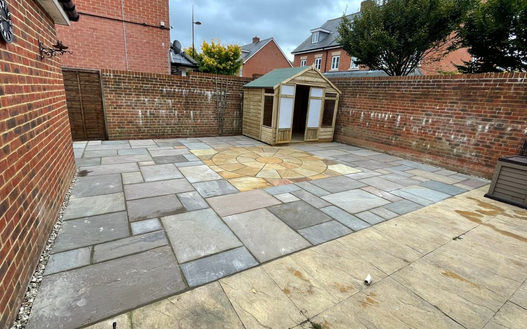 a newly laid patio with circular centre piece. There is a new small wooden summerhouse in one corner and large brick walls surrounding.