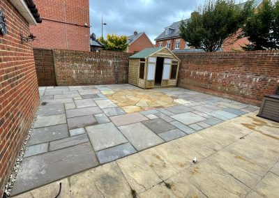 a newly laid patio with circular centre piece. There is a new small wooden summerhouse in one corner and large brick walls surrounding.