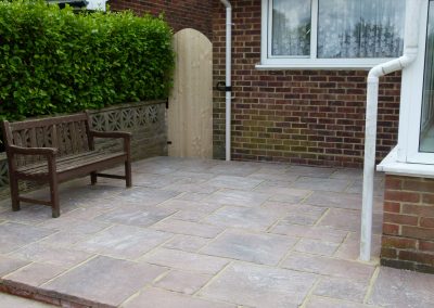a raised newly laid patio with a red tinge stone and a wooden bench and hedge to the side