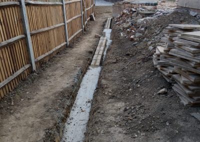 a side section of building area with drainage being laid. Pile of rubble and paving stones to one side and fence to the other