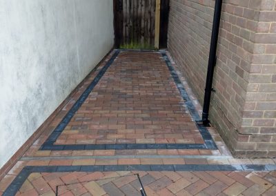 a small side path newly laid with red bricks and grey edging. The side walk has a white wall on one side, brick wall on other and a wooden gate