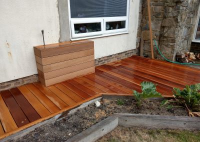 a small wooden deck area in corner of garden with borders in front