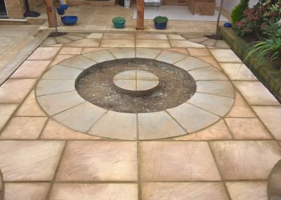 a circular paving area with light coloured slabs and space for planting. Garden border with green plants to the side