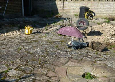 an old patio with upside down wheelbarrow, weeds and builders tools.